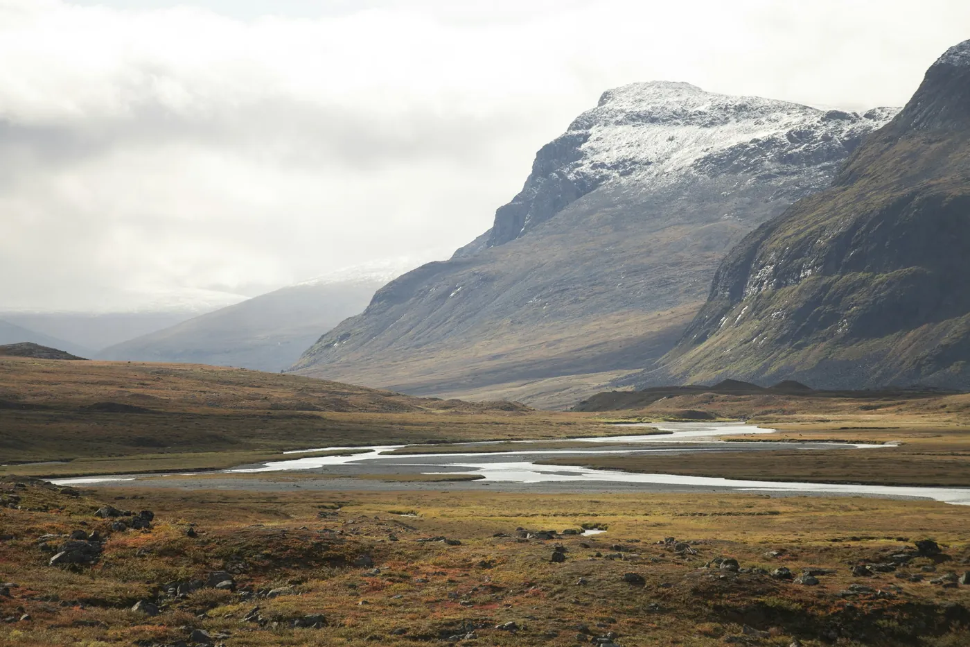 Tundra otoñal marrón y dorada con un río trenzado que lleva hacia montañas escarpadas con un ligero manto de nieve