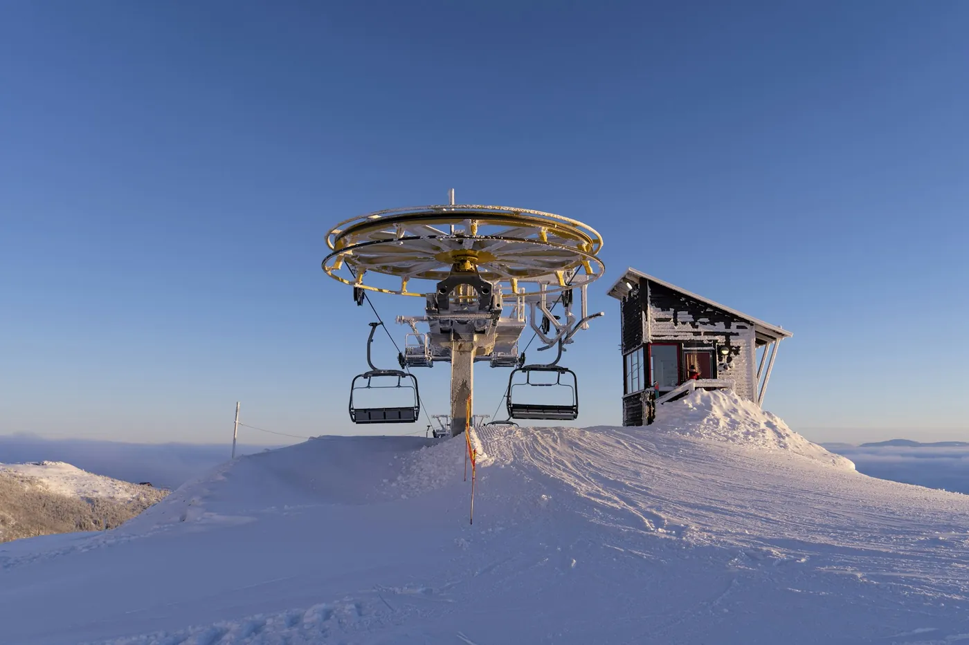Estación superior de un telesilla con sillas vacías y una cabaña pequeña en una cima nevada bajo un cielo azul despejado