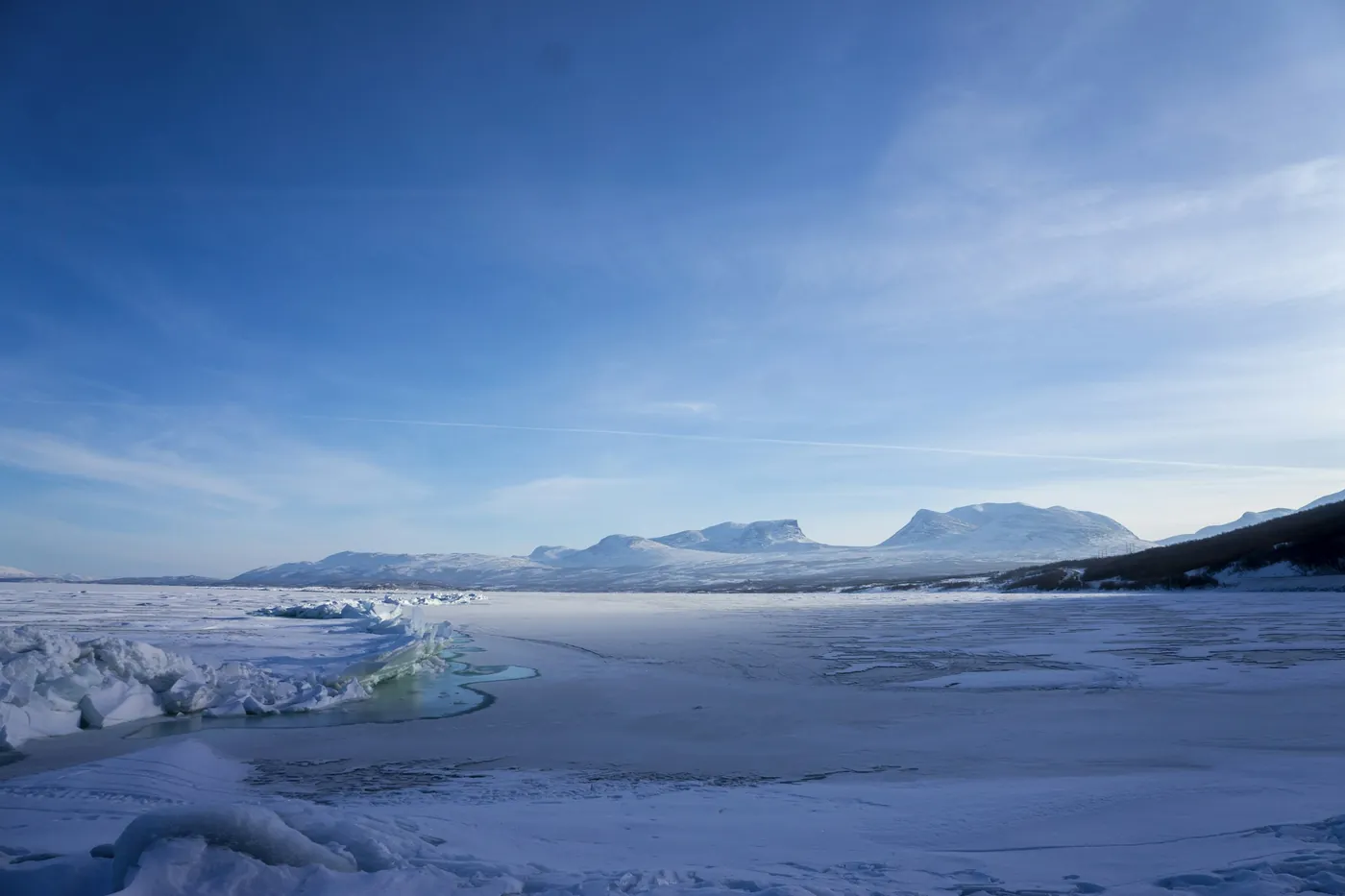 Lago helado cubierto de nieve, con crestas de hielo a lo largo de la orilla y una cordillera lejana bajo un cielo azul