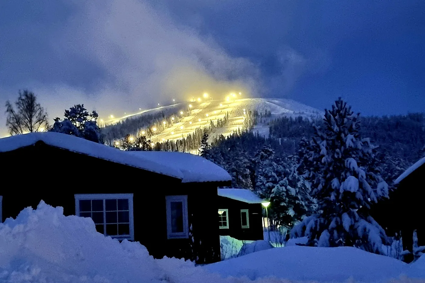 Pista da sci illuminata su una collina all’ora blu, cottage di legno scuro e abeti innevati in primo piano