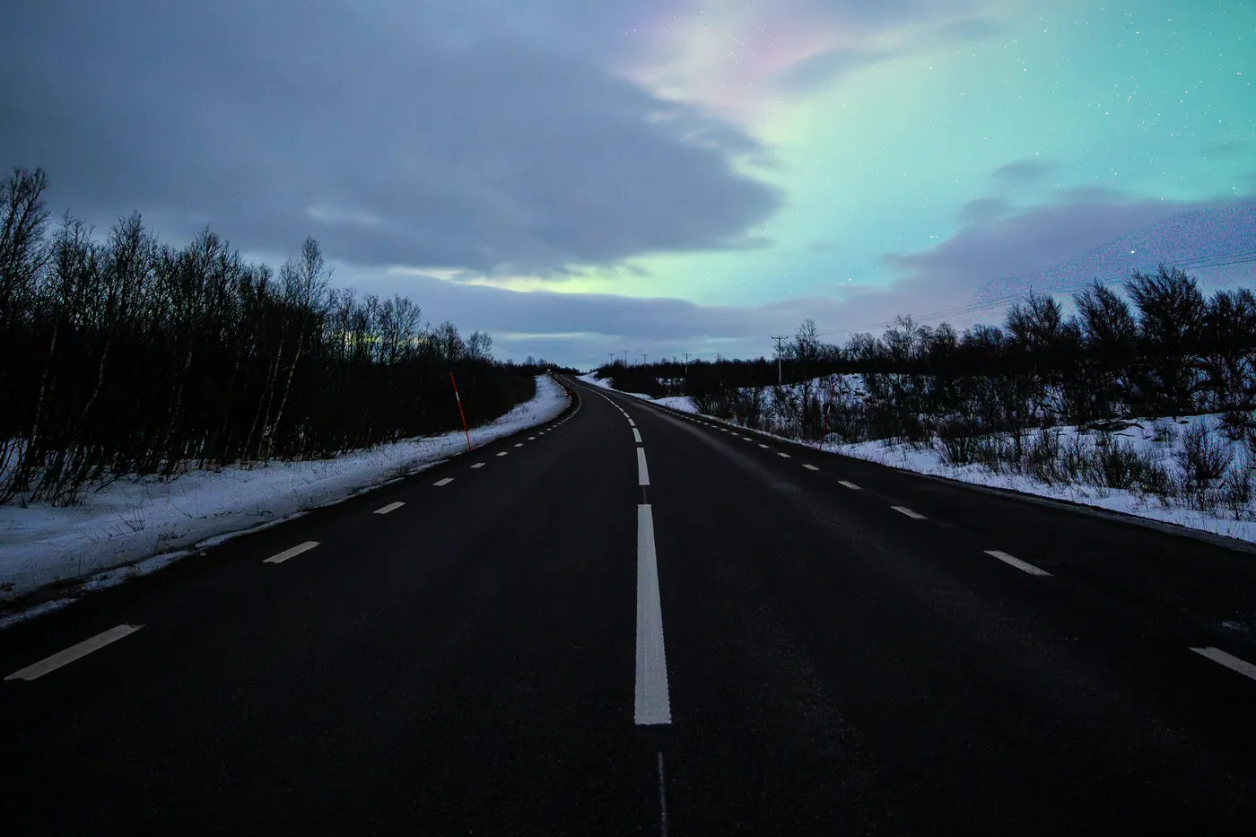 Carretera vacía de dos carriles entre matorral nevado de abedul, bajo una aurora y nubes de noche