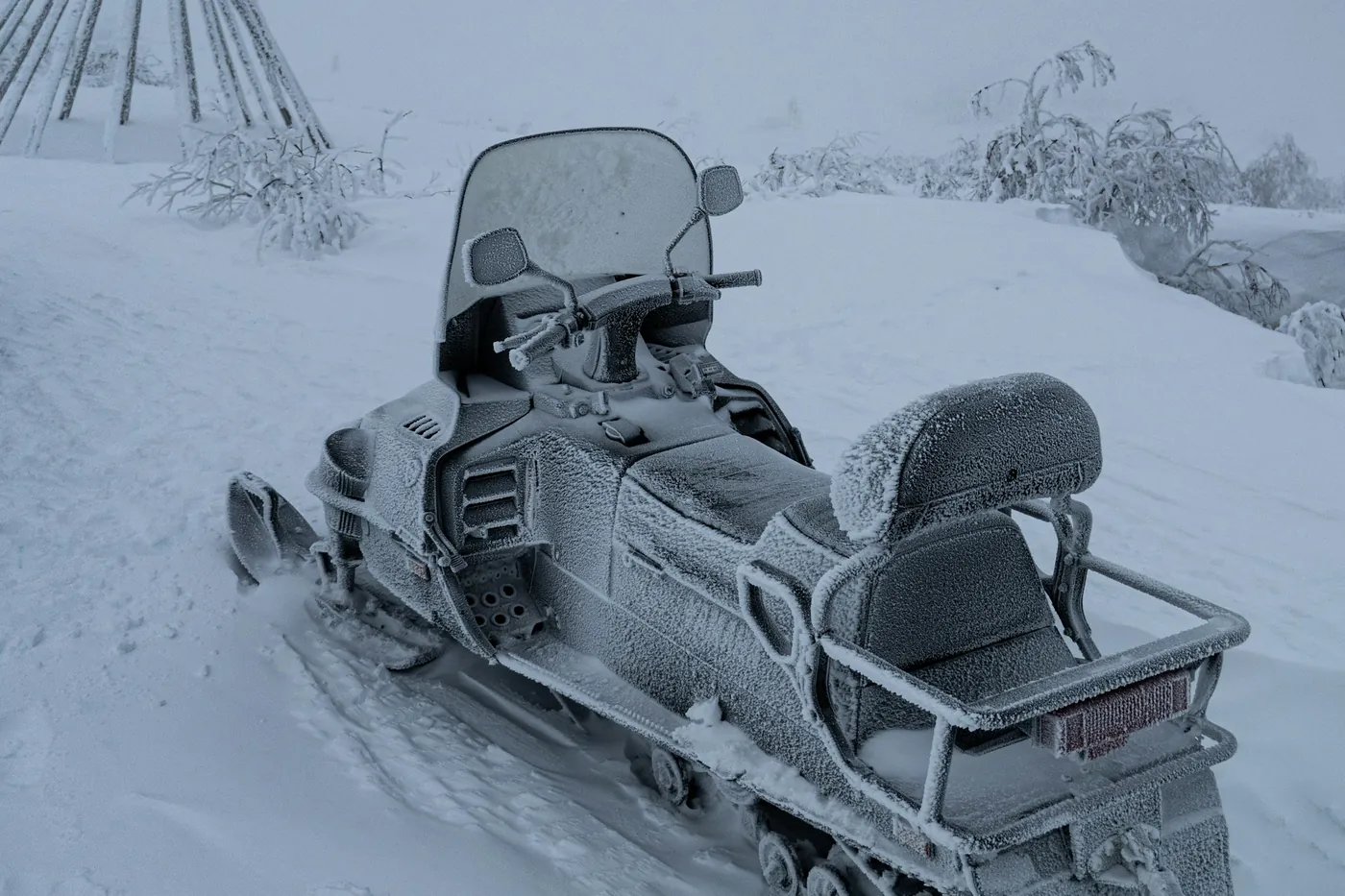 Moto de nieve de turismo cubierta de escarcha, aparcada en una montaña nevada entre la niebla, con los palos de la estructura de una lávvu detrás