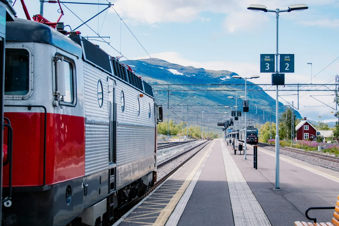 Locomotiva elettrica rossa e argento sul binario di una stazione, edificio di legno e montagna sullo sfondo, in estate